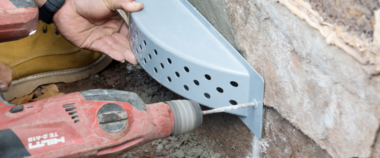 A person drilling a hole to attach a perforated metal piece to a concrete wall.