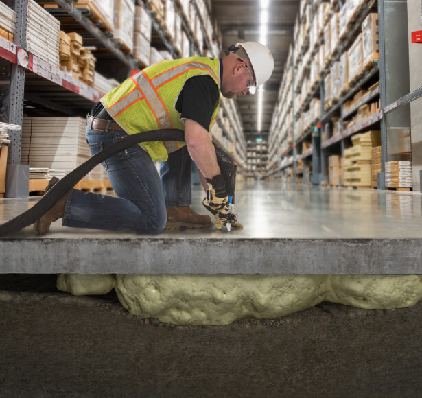 A worker injecting foam beneath a concrete slab in a warehouse to lift and stabilize it.